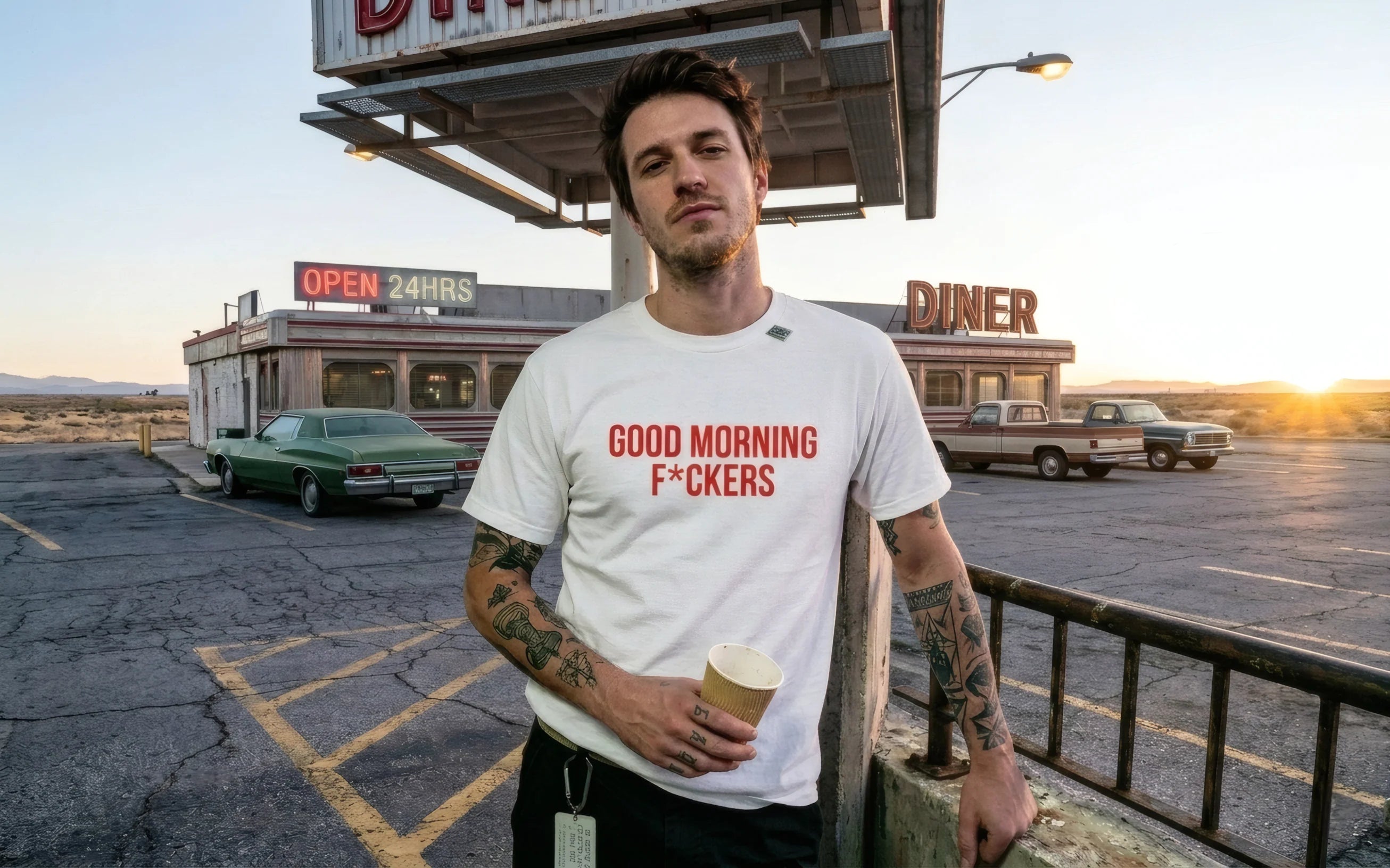 Mann mit Tattoos vor Retro-Diner am Morgen. Er trägt ein weißes Heavyweight Shirt mit rotem „GOOD MORNING F*CKERS“ Print und hält einen Kaffeebecher. Urbaner, stoischer Lifestyle-Shot im Empty Barrel Archive Stil. Staubiger Parkplatz, Sonnenaufgang.